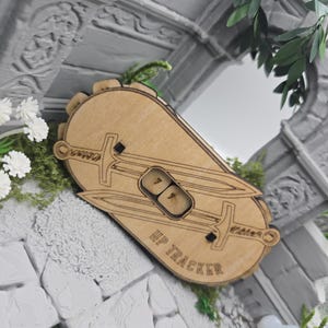 Könnte beinhalten: Ein hellbrauner HP-Tracker aus Holz in abgerundeter rechteckiger Form mit zwei eingravierten, gekreuzten Schwertern hinter einem zentralen Fenster, das die Zahl 77 anzeigt. Der Text HP TRACKER ist in die untere rechte Ecke eingraviert.