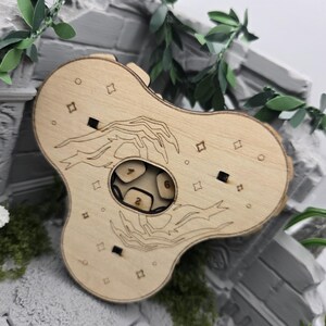 Könnte beinhalten: Eine hellfarbene Holzwürfelbox mit einer abgerundeten, dreilappigen Form. Die Oberfläche zeigt lasergravierte Illustrationen von zwei Händen und kleinen Sternsymbolen. Ein zentraler kreisförmiger Ausschnitt zeigt mehrere Würfel mit sichtbaren Zahlen.