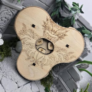 Könnte beinhalten: Ein hellfarbiger Würfelspinner aus Holz mit einer lasergravierten Drachenkopf-Grafik und Flammenmustern. In der kreisförmigen Mitte befinden sich kleine Holzwürfel mit sichtbaren Zahlen, die durch drei kleine schwarze Befestigungen gehalten werden.