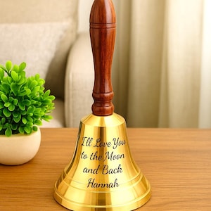 May include: A brass hand bell with a polished gold finish and a dark brown wooden handle topped with a small gold sphere. The bell features the engraved black text: "Ill Love You to the Moon and Back Hannah" centered on the front surface.