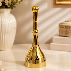 Puede incluir: Una campana de metal dorado pulido, alta y esbelta, con un mango alargado que presenta una parte superior esférica y un pomo decorativo central. La base de la campana es ancha y acampanada, con tres líneas horizontales grabadas en la parte inferior.