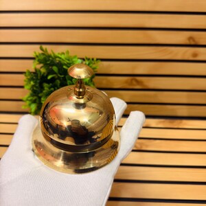 May include: A shiny brass bell with a rounded top and a circular base. The bell is held by a gloved hand. The background is a wooden wall with horizontal lines. The bell is a classic design, often used in hotels or reception areas.