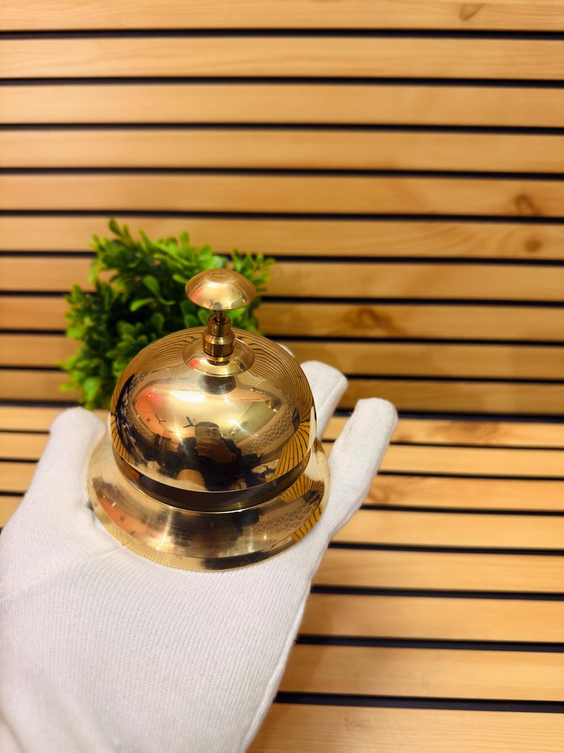May include: A shiny brass bell with a rounded top and a circular base. The bell is held by a gloved hand. The background is a wooden wall with horizontal lines. The bell is a classic design, often used in hotels or reception areas.