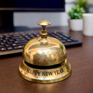May include: A shiny brass bell with a rounded top and a clapper. The base of the bell has the words "HAPPY NEW YEAR" engraved in a bold font. The bell is on a wooden surface, with a keyboard and monitor in the background.