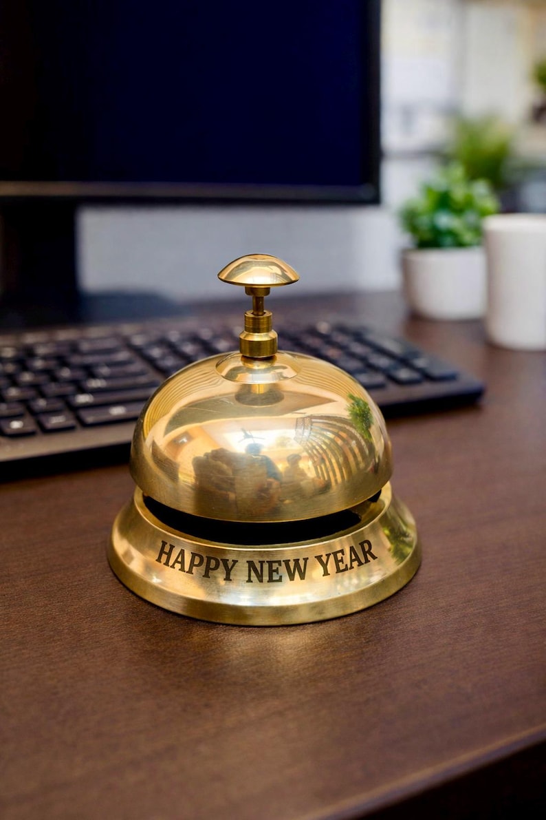 May include: A shiny brass bell with a rounded top and a clapper. The base of the bell has the words "HAPPY NEW YEAR" engraved in a bold font. The bell is on a wooden surface, with a keyboard and monitor in the background.