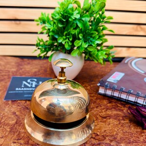 May include: A polished brass bell with a rounded top and a flared base. The bell has a small, round handle on top. A small green plant in a white pot is in the background.