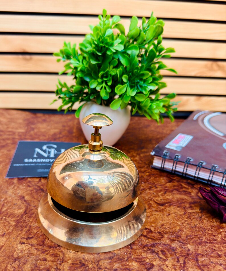 May include: A polished brass bell with a rounded top and a flared base. The bell has a small, round handle on top. A small green plant in a white pot is in the background.