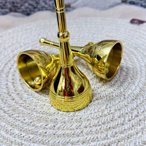 K&ouml;nnte beinhalten: Zwei gl&auml;nzende, goldfarbene Handglocken aus Metall mit langen, schlanken Griffen und ausgestellten Basen. Jede Glocke hat eine glatte, reflektierende Oberfl&auml;che mit feinen horizontalen Linien am Rand und einem runden Knopf am Griffende.