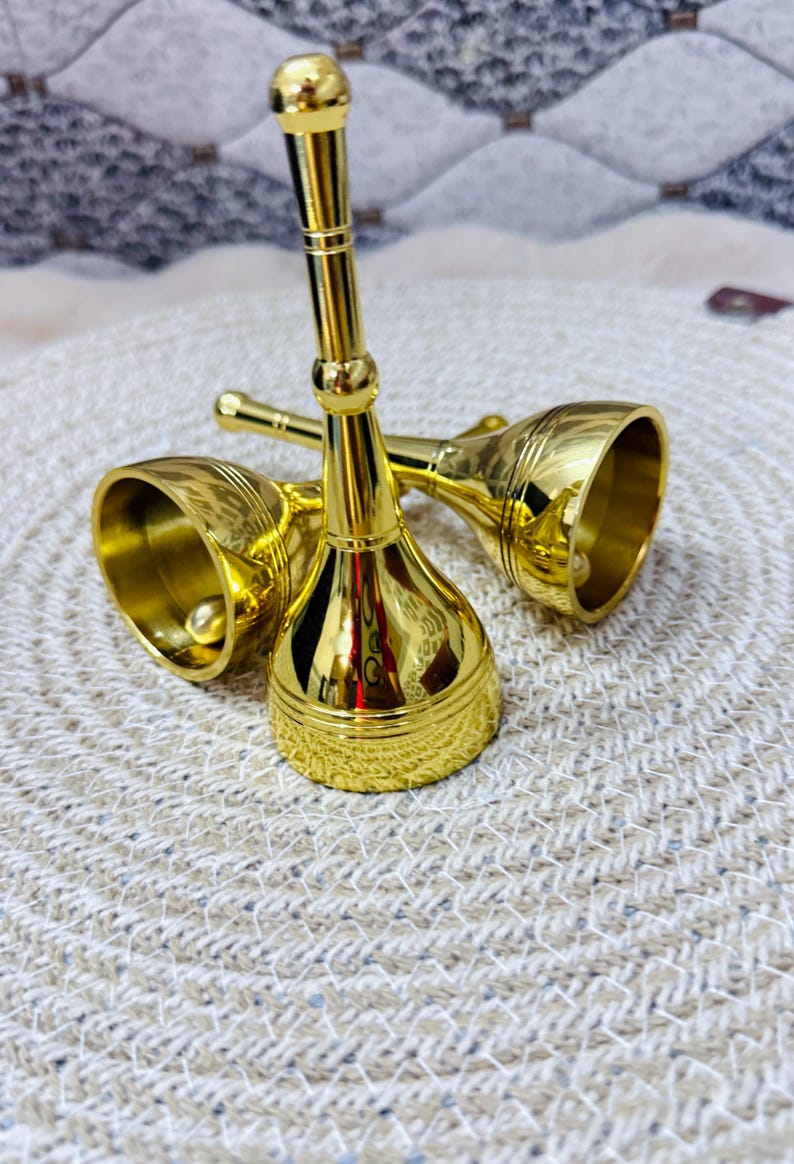 K&ouml;nnte beinhalten: Zwei gl&auml;nzende, goldfarbene Handglocken aus Metall mit langen, schlanken Griffen und ausgestellten Basen. Jede Glocke hat eine glatte, reflektierende Oberfl&auml;che mit feinen horizontalen Linien am Rand und einem runden Knopf am Griffende.