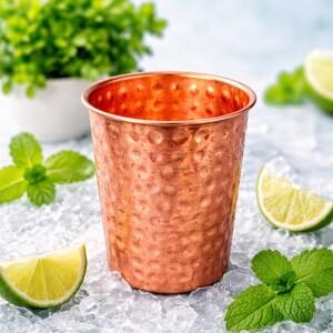 May include: A copper-colored, hammered-finish cup sits on crushed ice, with lime wedges and mint leaves. A small white pot with green plants is in the background. The cup is designed for cold beverages.