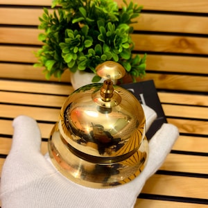 May include: A polished brass bell with a rounded base and a domed top. The bell has a detailed, ridged design and a small, round clapper. The bell is held in a white glove, with a green plant in the background.