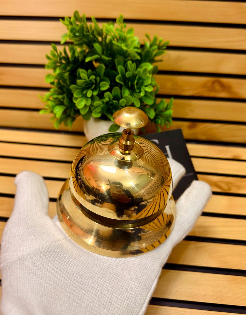 May include: A polished brass bell with a rounded base and a domed top. The bell has a detailed, ridged design and a small, round clapper. The bell is held in a white glove, with a green plant in the background.