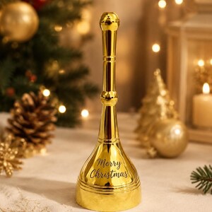 K&ouml;nnte beinhalten: Eine hohe, polierte goldfarbene Metallhandglocke mit einem langen, schlanken Griff und einem ausgestellten Fu&szlig;. Die Glocke tr&auml;gt den schwarzen Schriftzug Merry Christmas auf der Vorderseite und hat eine glatte, reflektierende Oberfl&auml;che.