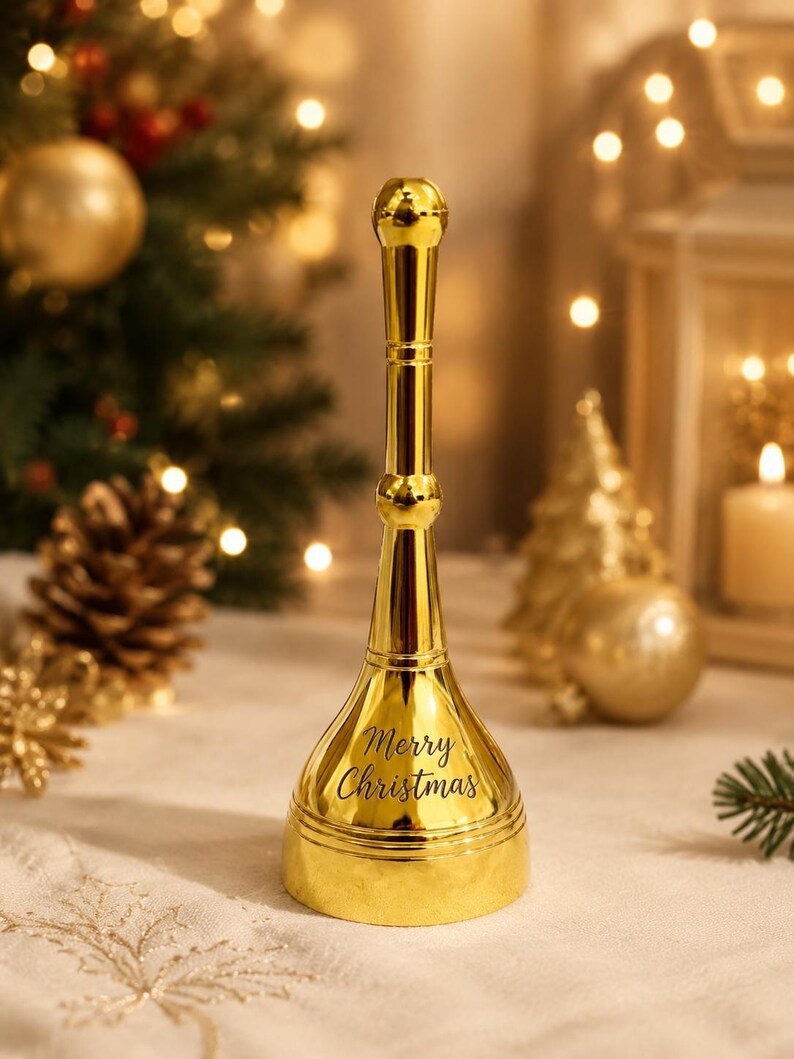 K&ouml;nnte beinhalten: Eine hohe, polierte goldfarbene Metallhandglocke mit einem langen, schlanken Griff und einem ausgestellten Fu&szlig;. Die Glocke tr&auml;gt den schwarzen Schriftzug Merry Christmas auf der Vorderseite und hat eine glatte, reflektierende Oberfl&auml;che.