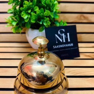 May include: A shiny brass bell with a rounded top and a small handle. The bell is sitting on a wooden surface. A small green plant in a white pot is in the background, along with a black card with the text "NH SAASNOVHUB".