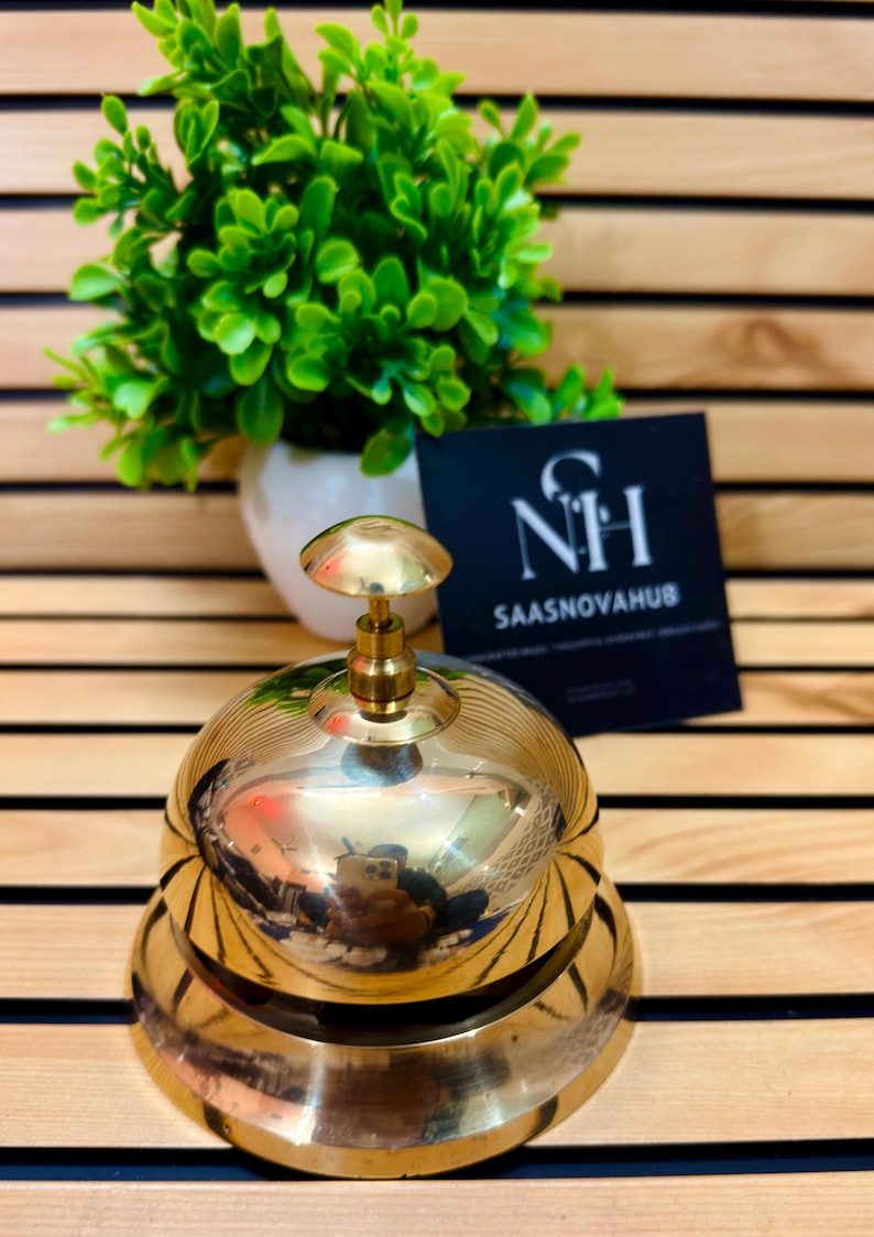 May include: A shiny brass bell with a rounded top and a small handle. The bell is sitting on a wooden surface. A small green plant in a white pot is in the background, along with a black card with the text "NH SAASNOVHUB".