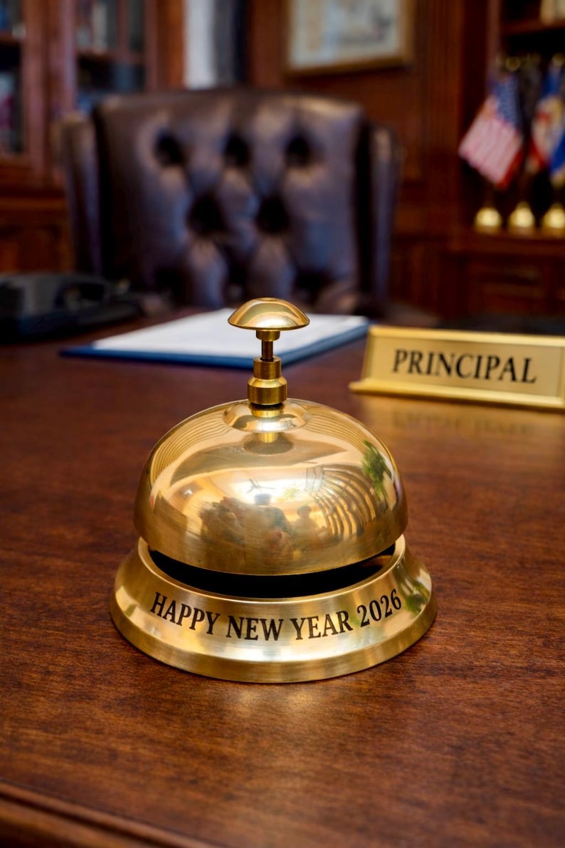 May include: A shiny gold-colored desk bell with a rounded top and a small handle. The base of the bell has the inscription "HAPPY NEW YEAR 2026" in black lettering. The bell sits on a dark wooden desk, with a "PRINCIPAL" sign in the background.