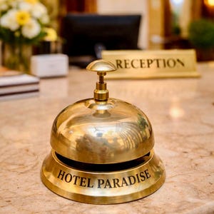 May include: A shiny brass hotel bell with the words "HOTEL PARADISE" engraved on the base. The bell sits on a marble countertop near a "RECEPTION" sign. The background is blurred, suggesting a hotel lobby setting.