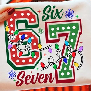 May include: A white t-shirt with a festive design featuring the words "Six" and "Seven" in green and red, with a string of colorful Christmas lights. The numbers "67" are also displayed.