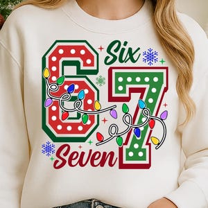 May include: Cream-colored sweatshirt with a festive design. The graphic features the number "67" in red and green, adorned with colorful string lights and the words "Six" and "Seven". Snowflakes add to the holiday theme.