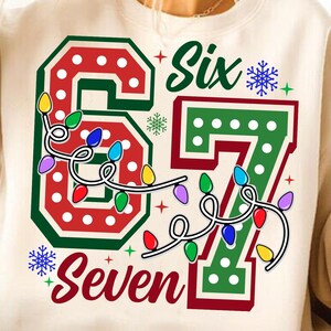May include: Cream-colored sweatshirt with a festive design. The graphic features the numbers "6" and "7" in red and green, adorned with string lights and the words "Six" and "Seven". Snowflakes and stars complete the holiday theme.