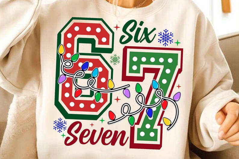 May include: Cream-colored sweatshirt with a festive design. The graphic features the numbers "6" and "7" in red and green, adorned with string lights and the words "Six" and "Seven". Snowflakes and stars complete the holiday theme.