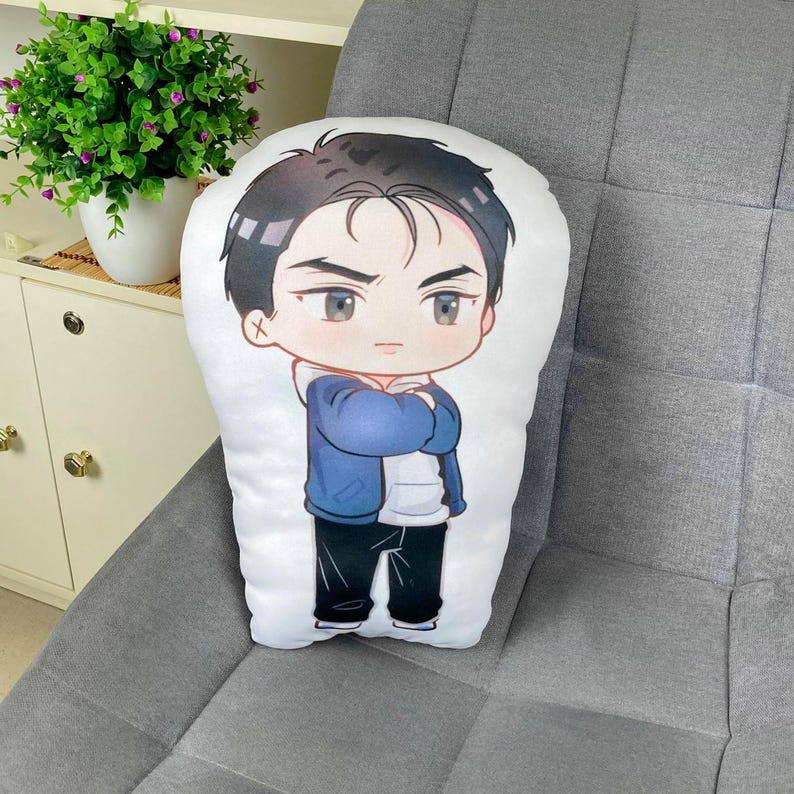 May include: A white pillow with a cartoon illustration of a male character. The character has black hair, gray eyes, and is wearing a blue jacket, white hoodie, and black pants. The pillow is resting on a gray upholstered surface.