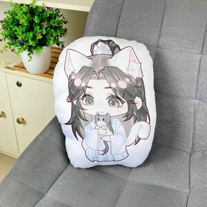 May include: White plush pillow featuring a cartoon illustration of a character with cat ears, holding a small gray and white cat. The character has long black hair and is wearing a light blue outfit. The pillow is on a gray upholstered surface.