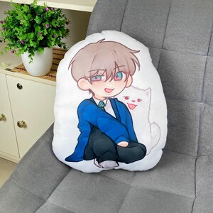 May include: White, oval-shaped pillow featuring a cartoon illustration of a young person in a blue blazer and black pants, with a white cat. The character has brown hair and is smiling. The pillow is on a gray upholstered surface.