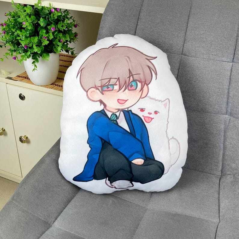 May include: White, oval-shaped pillow featuring a cartoon illustration of a young person in a blue blazer and black pants, with a white cat. The character has brown hair and is smiling. The pillow is on a gray upholstered surface.