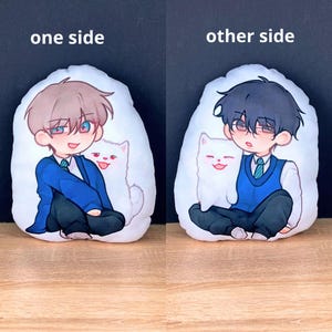 May include: Two white plush pillows with cartoon illustrations. One side features a boy with brown hair, blue eyes, and a white cat. The other side shows a boy with black hair, brown eyes, and a white cat. Text on top reads "one side" and "other side."