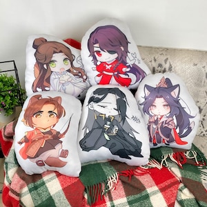 May include: A collection of six white, irregularly shaped throw pillows featuring anime-style character illustrations. The characters have various hair colors and wear different outfits, including red, black, and white. The pillows are arranged on a patterned blanket.