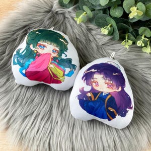May include: Two white plush pillows featuring anime-style character illustrations. The left pillow shows a character with green hair in a pink and blue outfit. The right pillow shows a character with purple hair in a dark blue outfit.