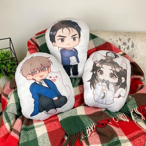 Puede incluir: Tres cojines blancos con ilustraciones de personajes de estilo anime de dibujos animados. Un cojín presenta un personaje con una chaqueta azul, otro con un traje azul y el tercero con orejas de gato sosteniendo un gato. Los cojines están sobre una manta a cuadros.