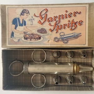 Könnte beinhalten: Ein Set aus einer Gebäck-Spritze aus Metall in einem Karton. Der Deckel zeigt eine Illustration einer Person, die einen Kuchen verziert, mit dem Text Garnier-Spritze. Das Set enthält eine silberfarbene Spritze mit Ringgriffen und sechs Tüllen.