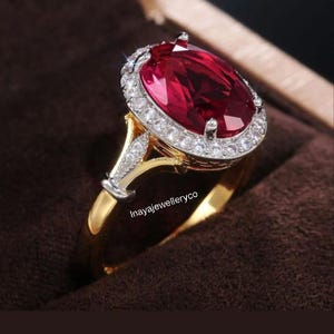 May include: A gold ring featuring a large, oval-cut, deep red gemstone, encircled by a halo of smaller, clear stones. The ring's band is gold, with a split shank design. The ring is set against a brown backdrop.