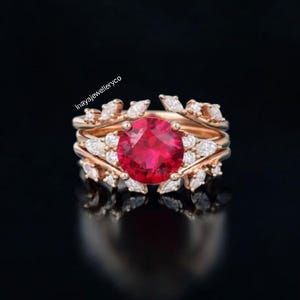 May include: A rose gold ring set featuring a large, round, red gemstone as the centerpiece. The ring is surrounded by smaller, clear gemstones and decorative leaf-like accents. The background is black, highlighting the ring's details.