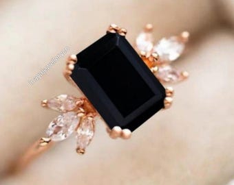 Könnte beinhalten: Roségoldener Ring mit einem großen, rechteckigen schwarzen Edelstein im Zentrum. Der Ring ist mit kleineren, klaren Edelsteinen verziert. Das Band ist schmal und das Design elegant und modern. Ein Schmuckstück.