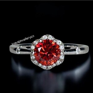 May include: A silver ring with a large, round, red gemstone at the center, surrounded by a halo of smaller, clear stones. The ring band has a simple design with small, clear stones. The text "Inayajewelleryco" is visible.