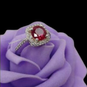May include: A silver ring with a large, round, red gemstone at the center, surrounded by smaller, clear stones. The ring is set on a purple rose, with the text "Inayajewelleryco" visible on the ring's band. The background is black.