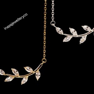 May include: Two necklaces with leaf-shaped pendants. One necklace is gold-toned, the other is silver-toned. Each pendant features clear, marquise-cut stones. The necklaces hang against a black background. The text "inayajewelleryco" is visible.