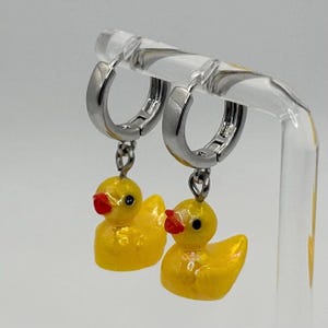Puede incluir: Pendientes de aro plateados con colgantes de pato amarillo. Los patos tienen picos rojos y ojos negros. Los pendientes se muestran sobre un soporte transparente sobre un fondo blanco.