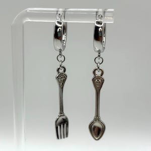 Puede incluir: Pendientes plateados con dijes de tenedor y cuchara. Los dijes cuelgan de aros. El tenedor tiene cuatro púas y la cuchara tiene un cuenco redondeado. Los pendientes se muestran sobre un fondo blanco.