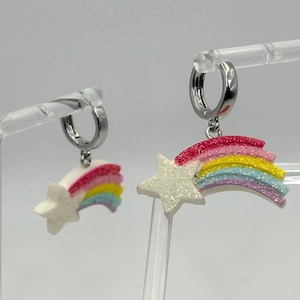 May include: A pair of silver hoop earrings. Each earring features a white glittery star and a rainbow-coloured arc. The earrings are displayed on clear plastic stands against a white background. The earrings have a shooting star design.