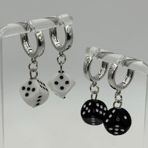 May include: Silver hoop earrings with dice charms. One pair features white dice with black dots, the other pair has black dice with white dots. The earrings are displayed on a clear stand against a white background.