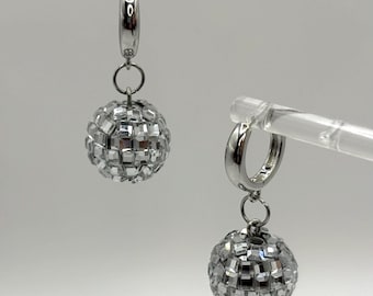 May include: A pair of silver-toned hoop earrings. Each earring features a small, round, disco ball charm. The disco ball charms are made of many small, reflective, square-shaped pieces. The earrings are displayed against a white background.