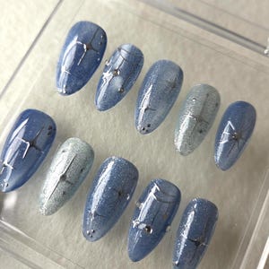 May include: A set of ten artificial nails in a clear plastic case. The nails are oval-shaped with a blue glitter base and silver accents. Some nails have small, silver embellishments.