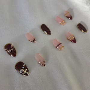 Può includere: Una collezione di unghie finte a forma di mandorla in vari design. Alcune unghie sono marrone scuro, altre rosa pallido. Gli abbellimenti includono catene d'oro, croci e piccole perle. Alcune unghie hanno un design maculato.