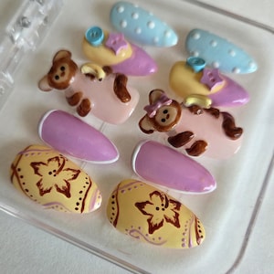 May include: A set of ten press-on nails in a clear plastic case. The designs include light blue nails with white dots, yellow nails with blue and purple buttons, pink nails with brown teddy bears, solid purple nails, and yellow nails with brown floral patterns.