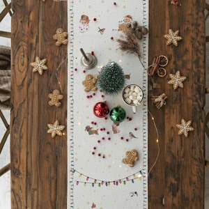 May include: A Christmas-themed table runner featuring dogs, snowmen, and Christmas trees. The runner is adorned with gingerbread biscuits, baubles, and a small Christmas tree. The table is made of dark wood.